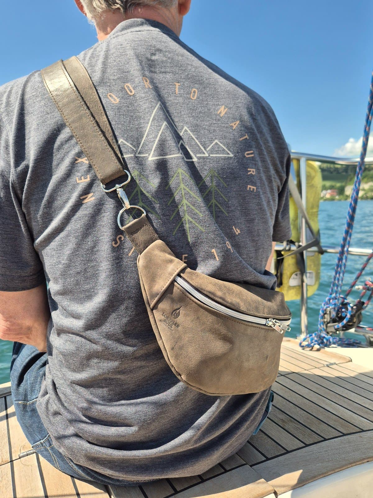 Mann sitzt auf einem Segelboot und trägt die Sling in Taupe quer über dem Rücken